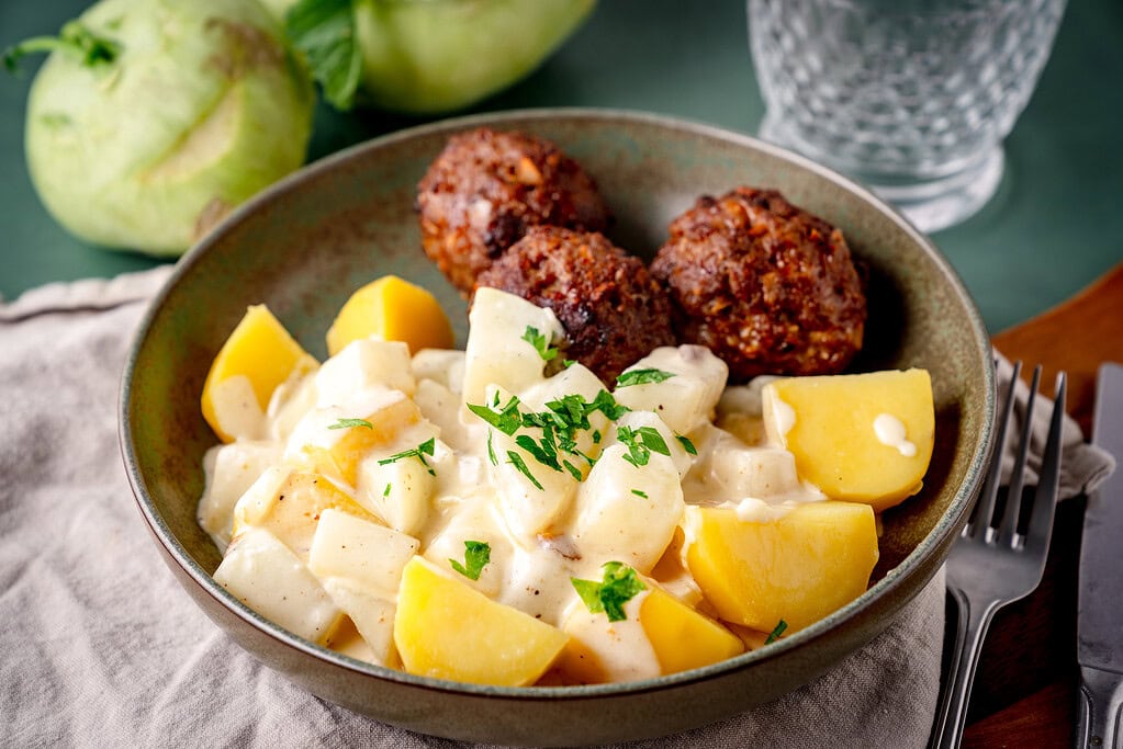 Creamed kohlrabi with boiled potatoes and meatballs on a green plate on a green background.