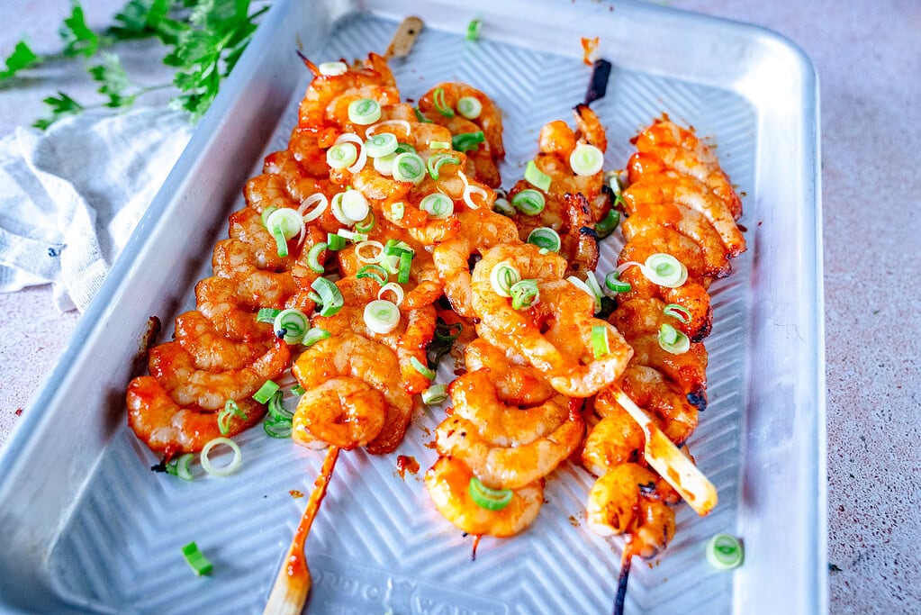 Five gochujang shrimp skewers topped with spring onions on a tray