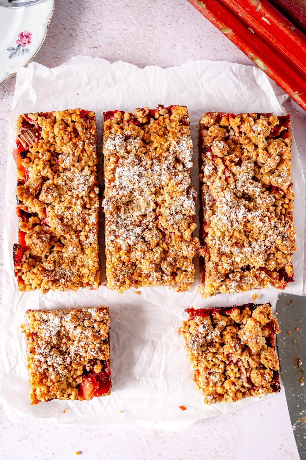 Rhubarb crumble cake Topview cut into pieces on baking paper surrounded by rhubarb sticks