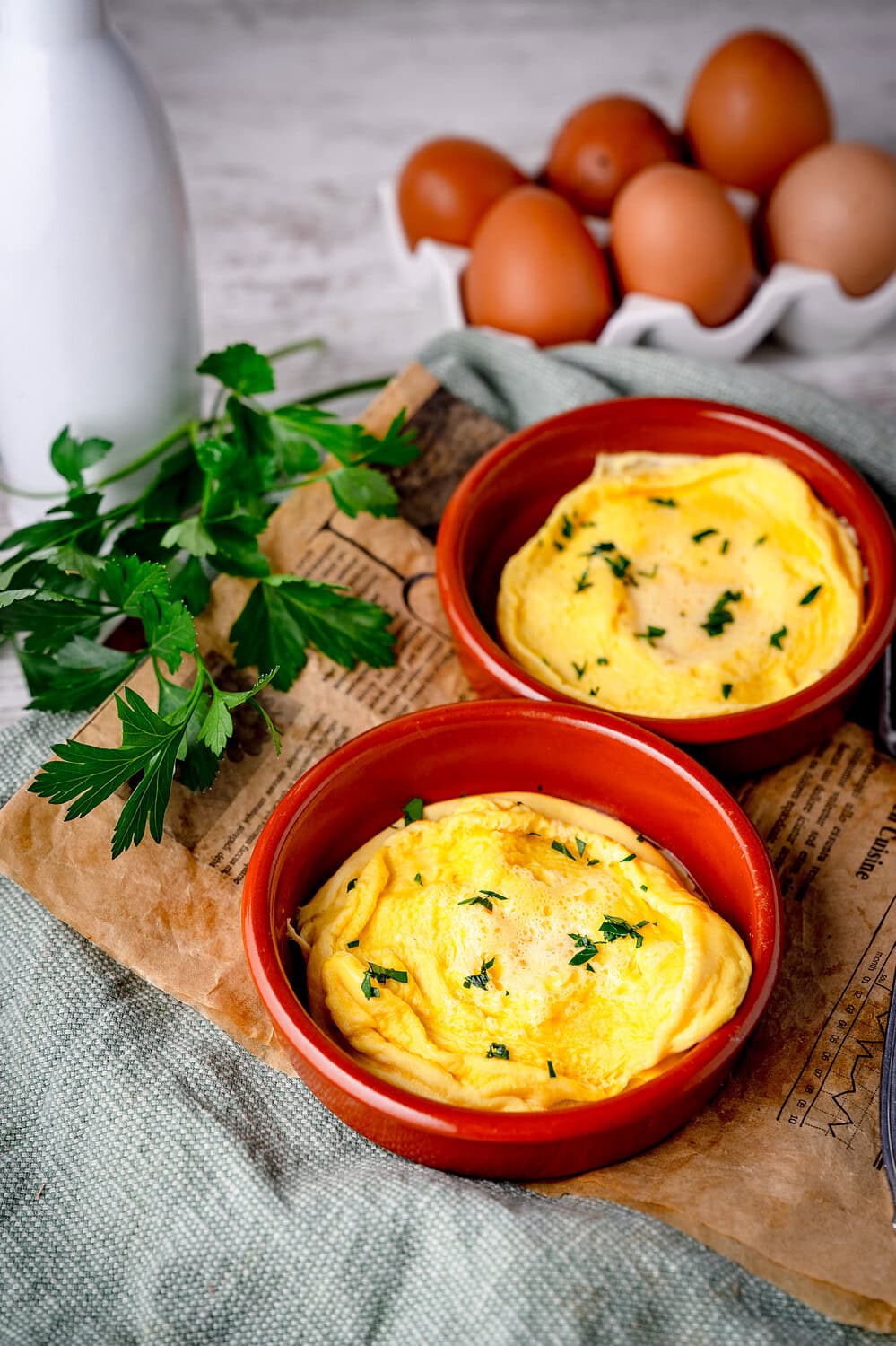 Frisches Omelett in roten Schälchen mit Kräutern garniert. Im Hintergrund sieht man Eier und Petersilie