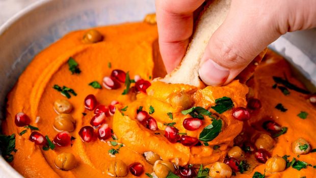 Roter Paprikahummus mit Kichererbsen, Granatapfel und Petersilie, eine Hand, ein Stück Brot, das sich etwas vom Dipp nimmt