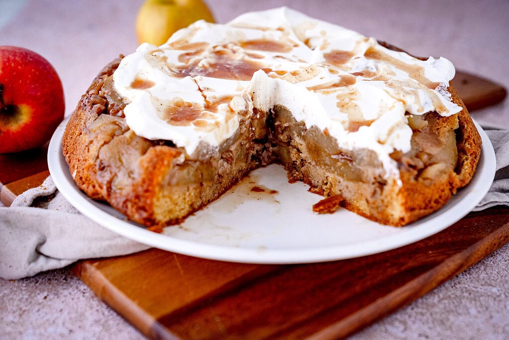Gestürzter Apfelkuchen mit Walnüssen und Sahne sowie Gewürzsud auf weißem Teller mit Äpfeln