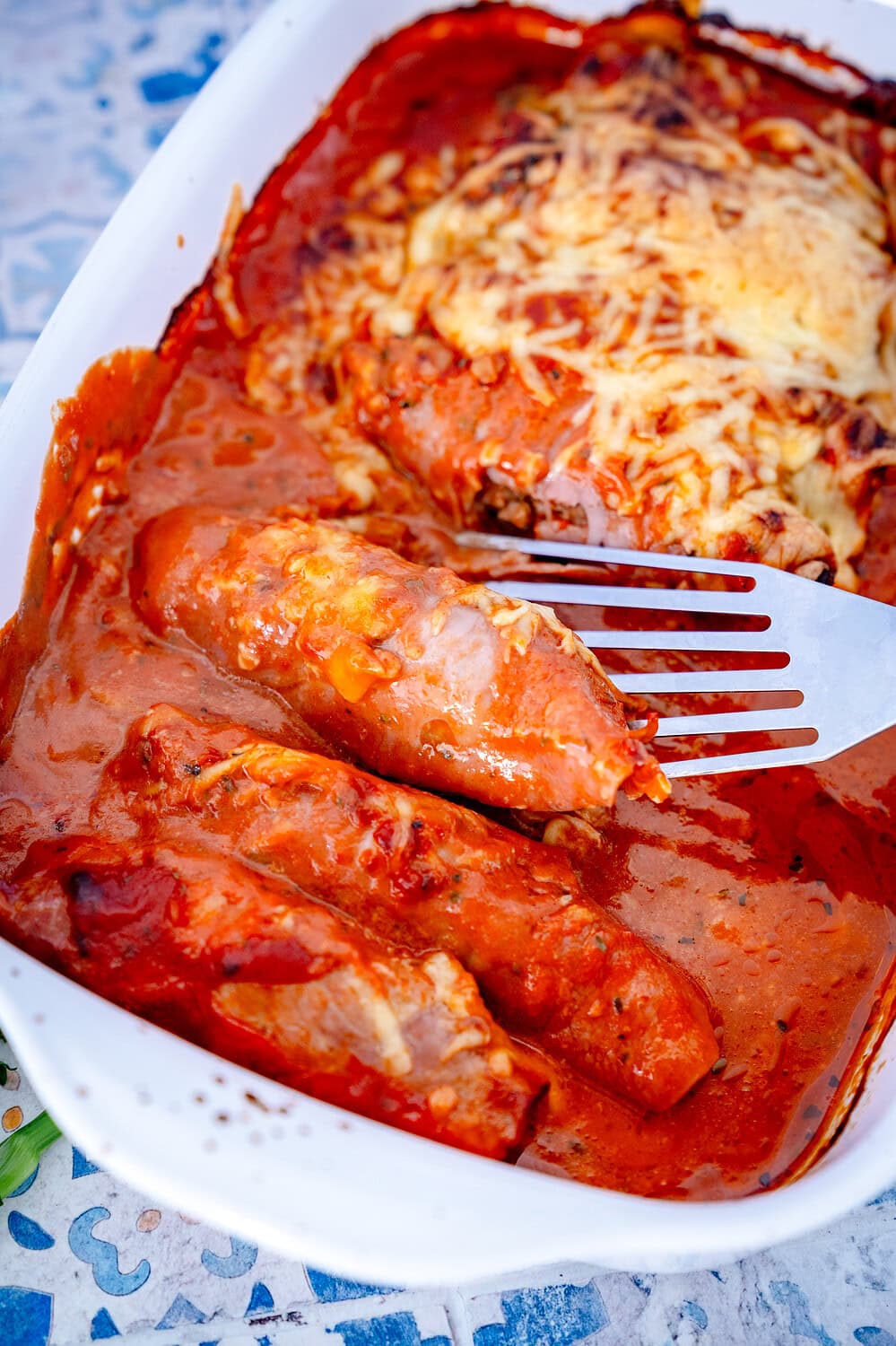 Baked ham meat rolls in a baking dish with a spatula, just removed from the tin. Red tomato cream sauce and melted cheese can be seen.
