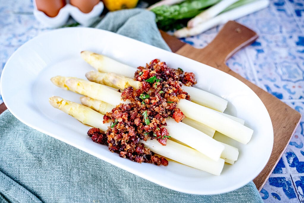 An oblong white plate on a wooden board with several spears of asparagus and bacon sauce