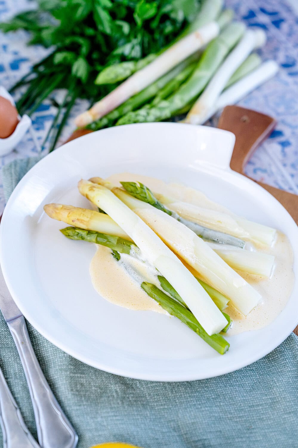 white and green asparagus on a white plate with a flash of hollandaise. Herbs and fresh asparagus can be seen in the background.