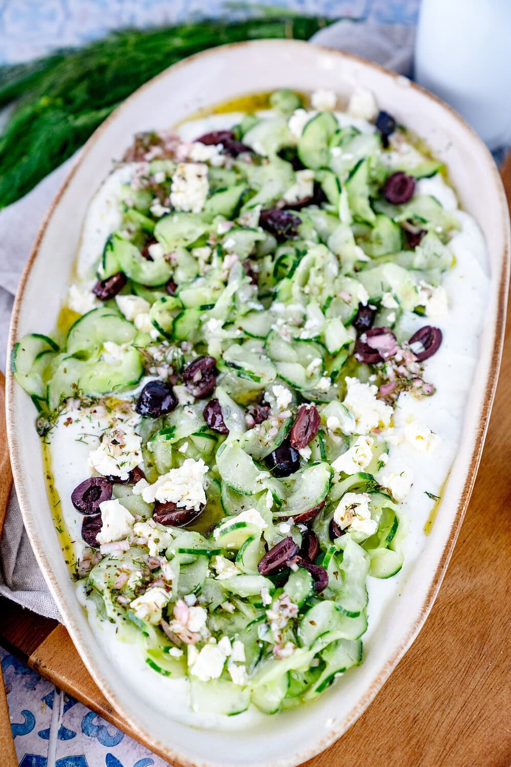 Draufsicht auf den griechischen Gurkensalat, der in einer großen ovalen Servierform liegt und mit Fetakrümeln und Oliven bedeckt ist