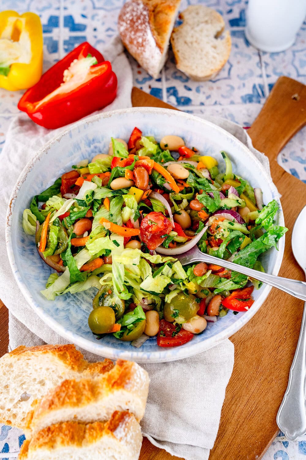 Mediterranean bean salad on a deep blue plate with a spoon in it. The plate is on a wooden board, with bread and sliced ​​peppers lying around it