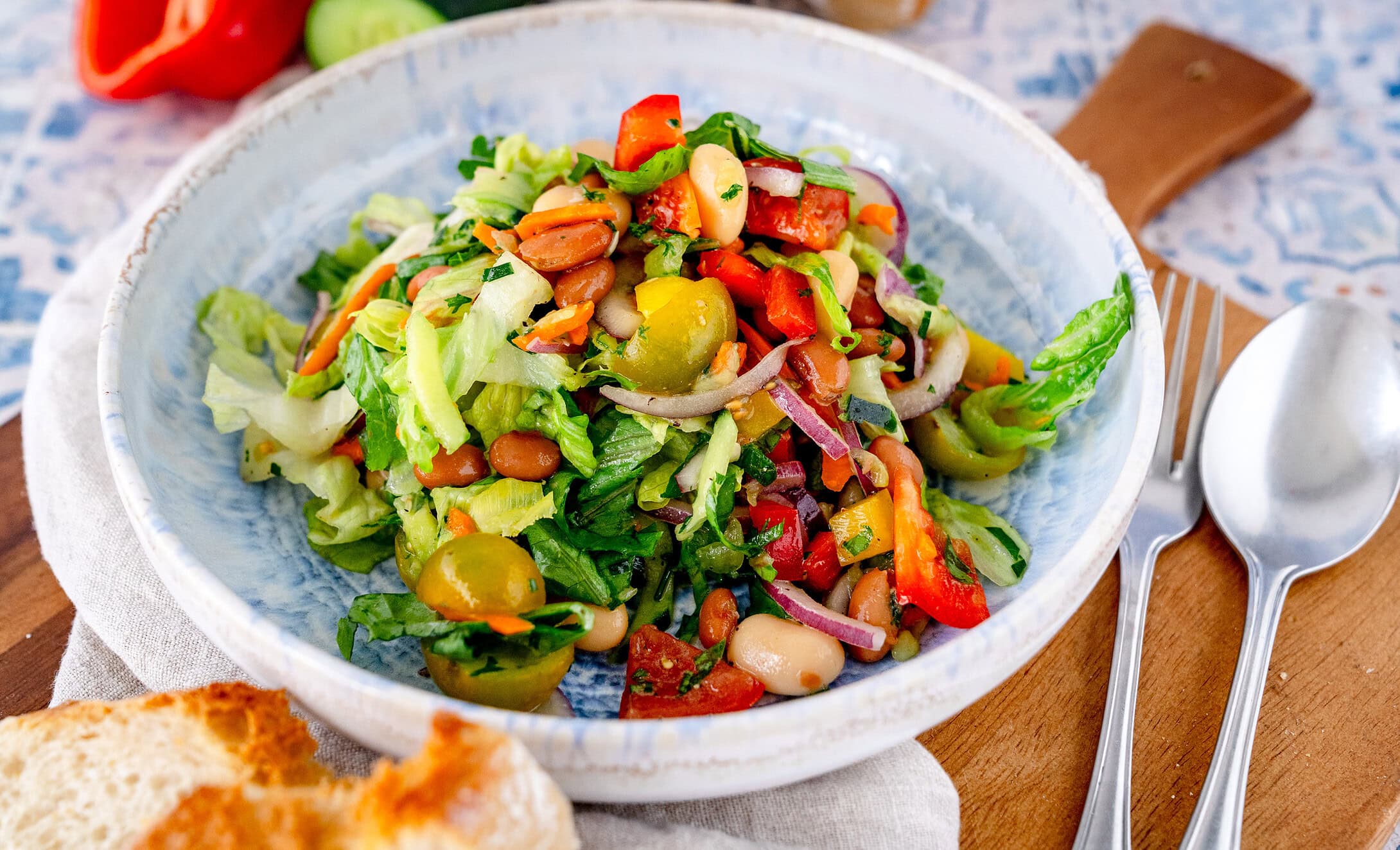 Mediterraner Bohnensalat in einem tiefen blauen Teller auf einem Holzbrett mit Besteck daneben
