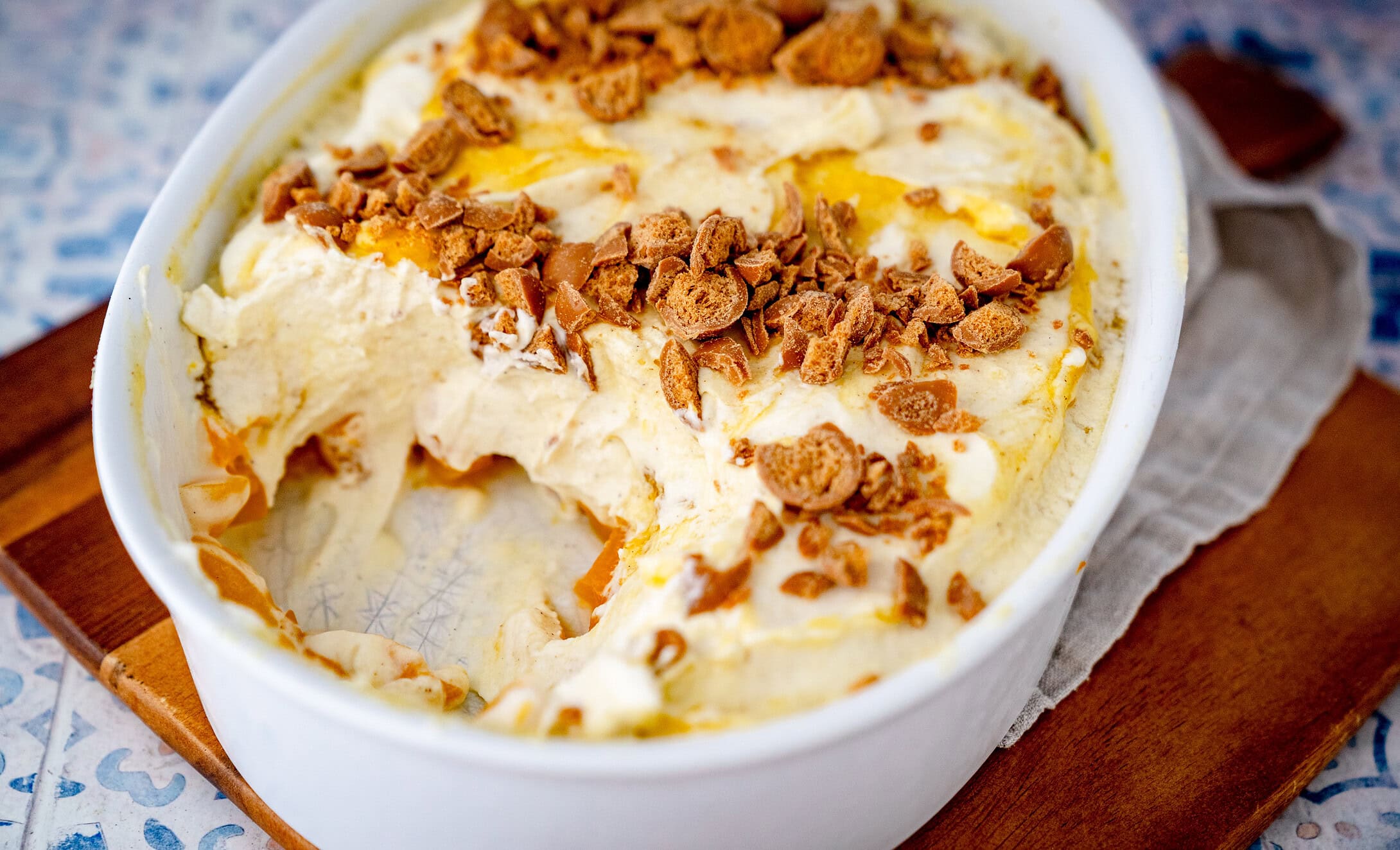 Solero Dessert mit Maracuja Quark in einer Auflaufform aus der bereits ein Stück entnommen wurde. Man sieht in der untersten Schicht gewürfelte Pfirsiche, darauf eine Sahne-Quark-Creme mit gelber Sauce und Keksbröseln.
