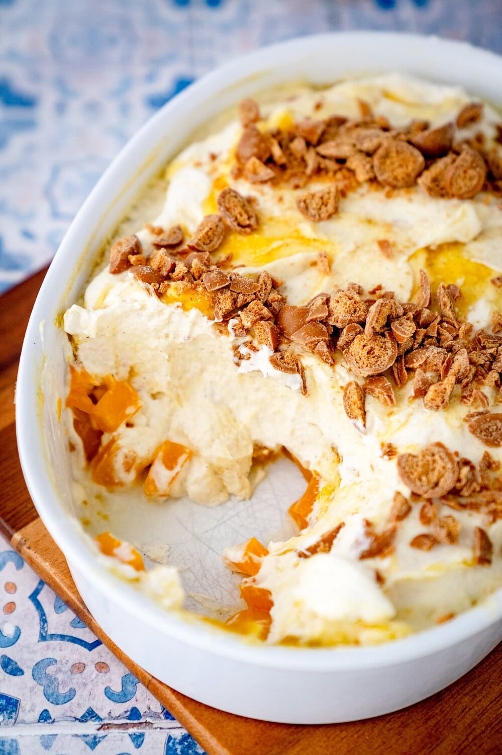 Solero dessert with passion fruit quark in a baking dish from which a piece has already been removed. You can see diced peaches in the bottom layer, followed by a cream quark cream with yellow sauce and biscuit crumbs.