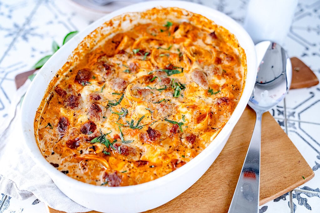 Tortellinigratin in einer weißen Auflaufform auf einem Holzbrett mit einem großen Servierlöffel daneben