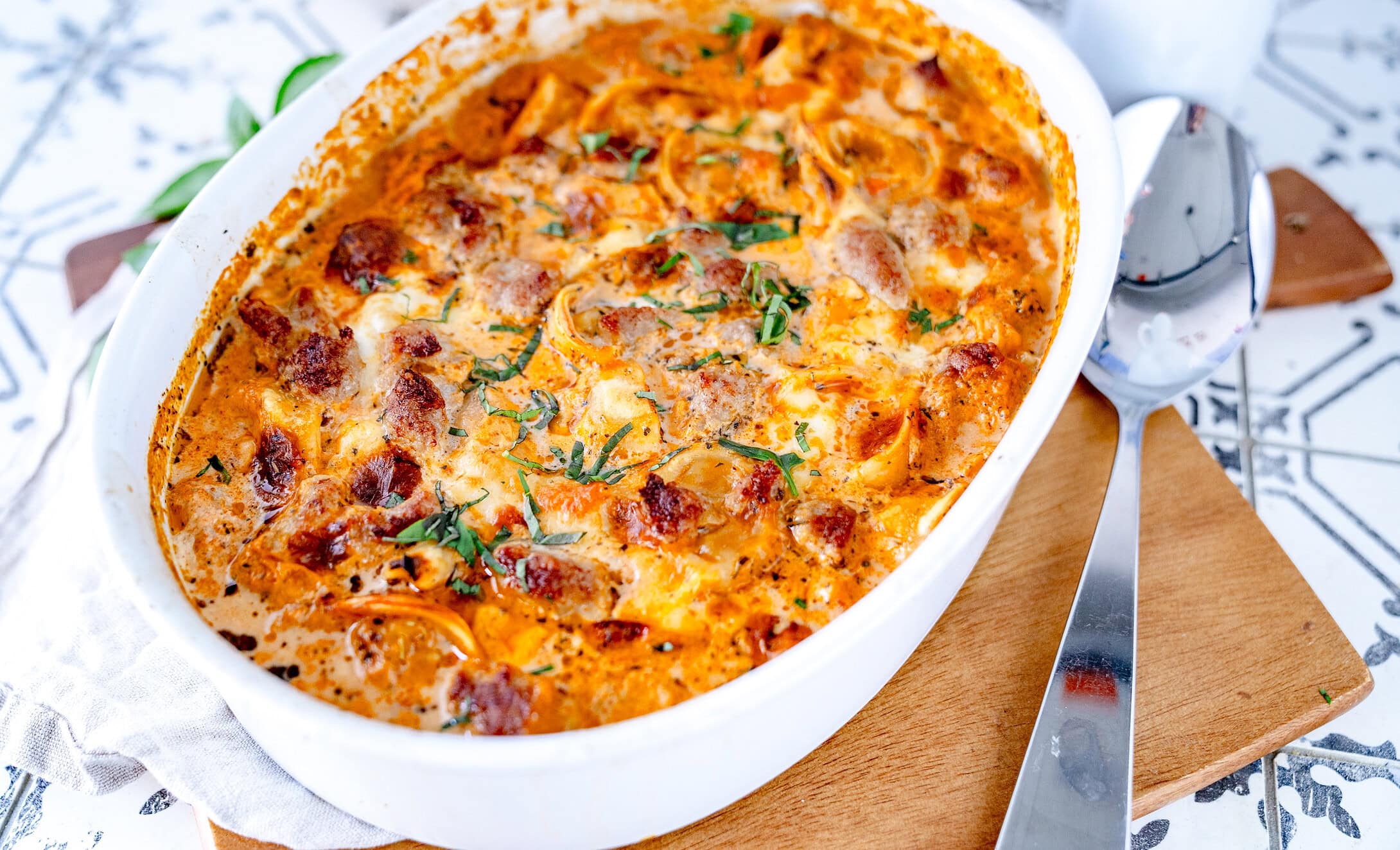 Tortellinigratin in einer weißen Auflaufform auf einem Holzbrett mit einem großen Servierlöffel daneben