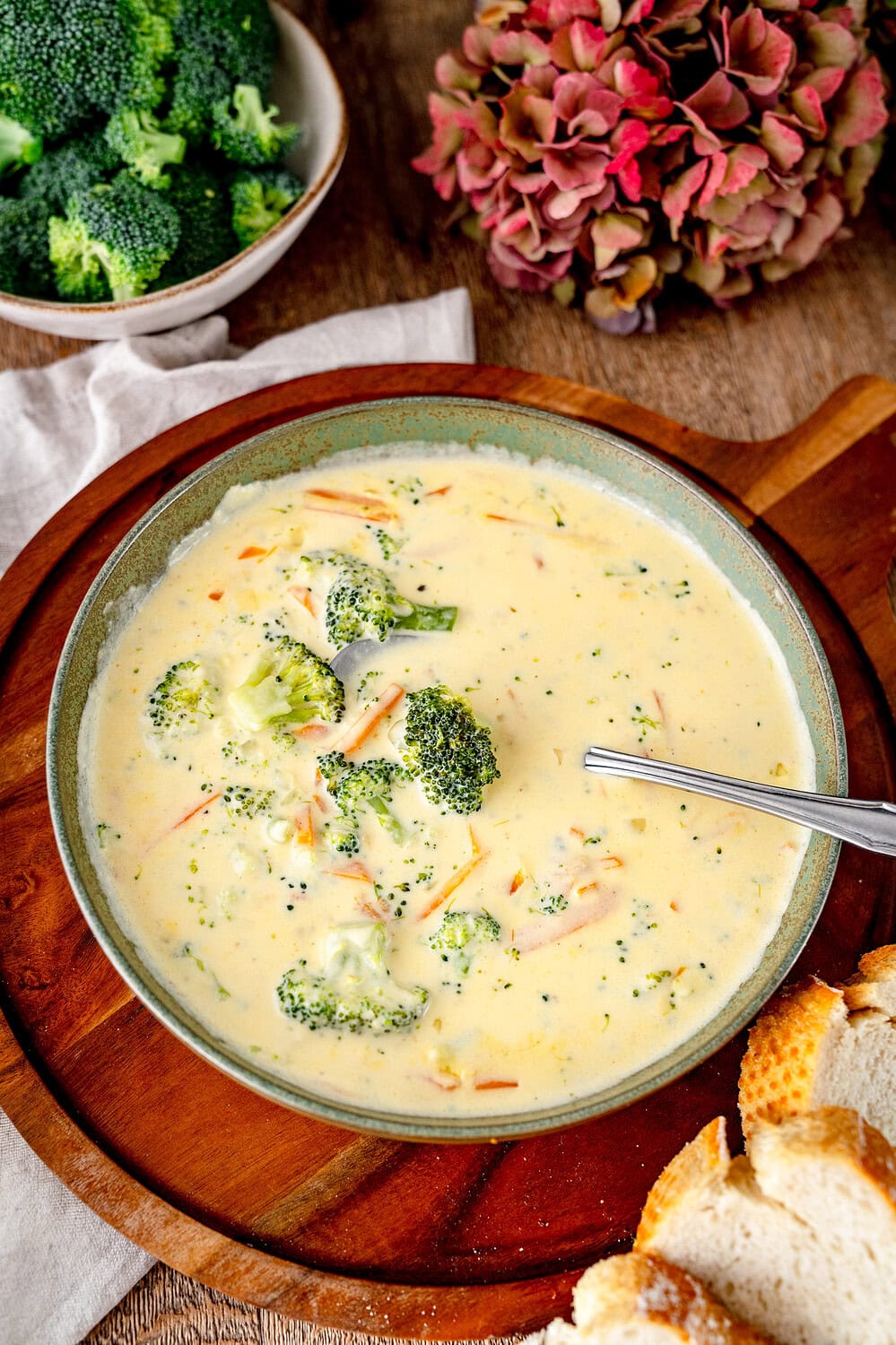 Brokkoli-Cheddar-Suppe in einem grünen tiefen Teller mit einem Löffel in der Suppe. Der Teller steht auf einem Holzbrett und Baguette befindet sich drumherum.