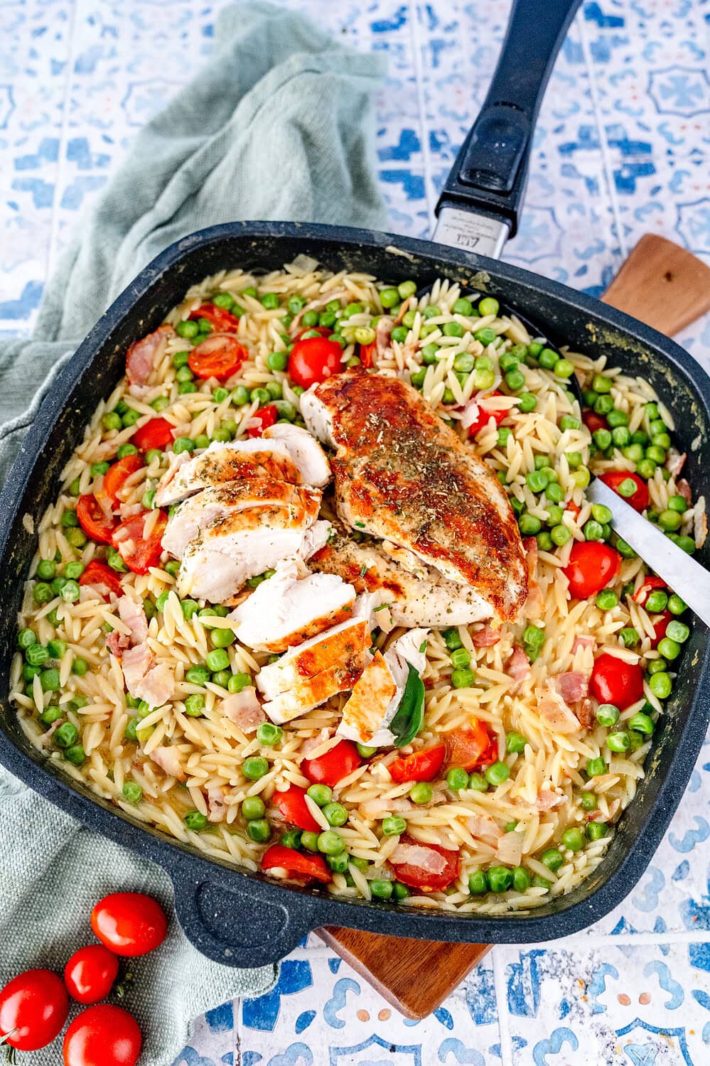 Topview auf eine Pfanne mit Orzo Nudeln, Erbsen, Tomaten und Bacon. Ein gegartes Hähnchenbrustfilet liegt obendrauf und ist schon in Stücke geschnitten. Ein Servierlöffel liegt in der Pfanne.