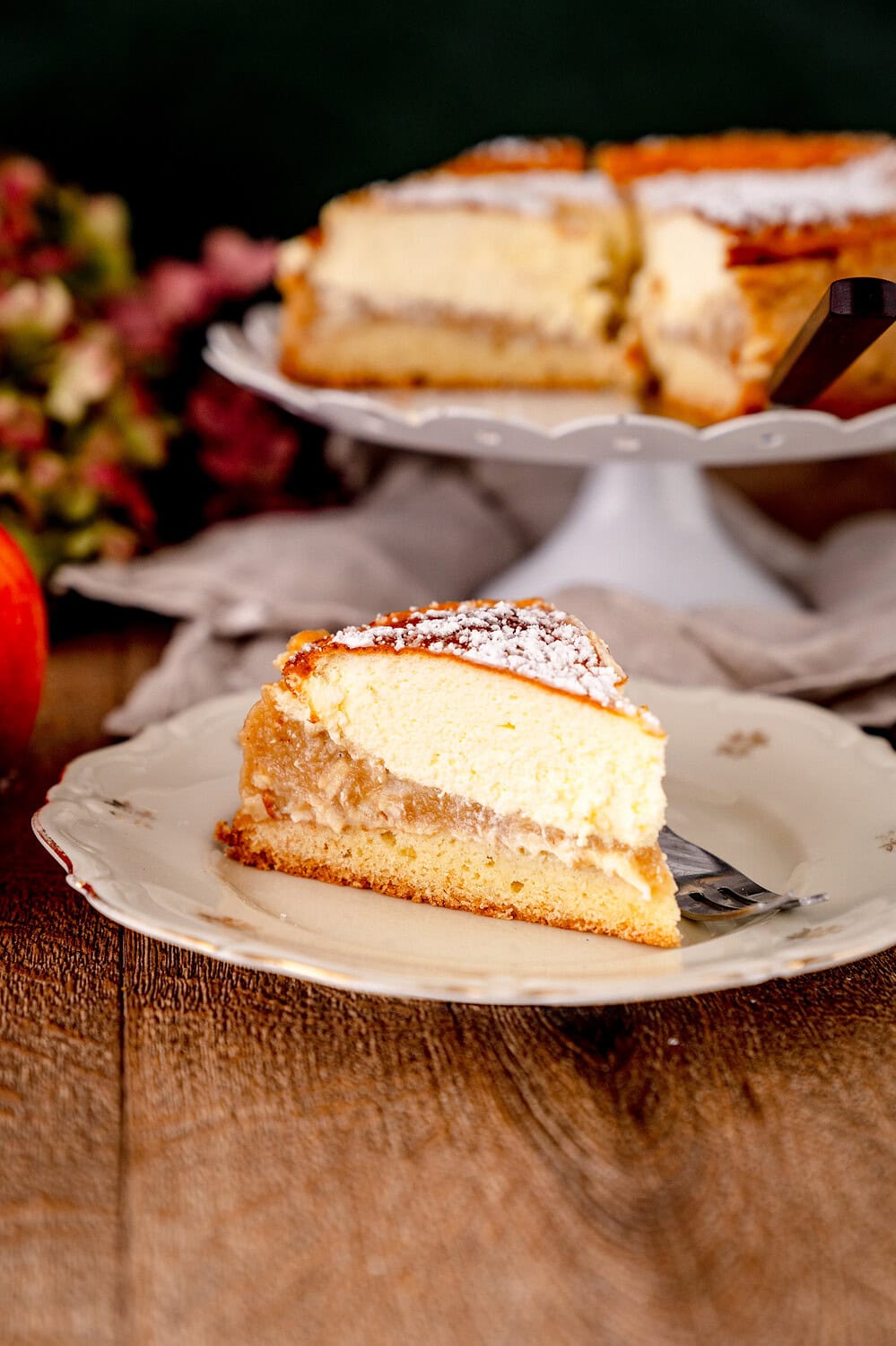 Ein stück Apfelmus-Schmandkuchen auf einem geblümten Teller mit einer Gabel daneben, im Hintergrund steht der ganze Kuchen