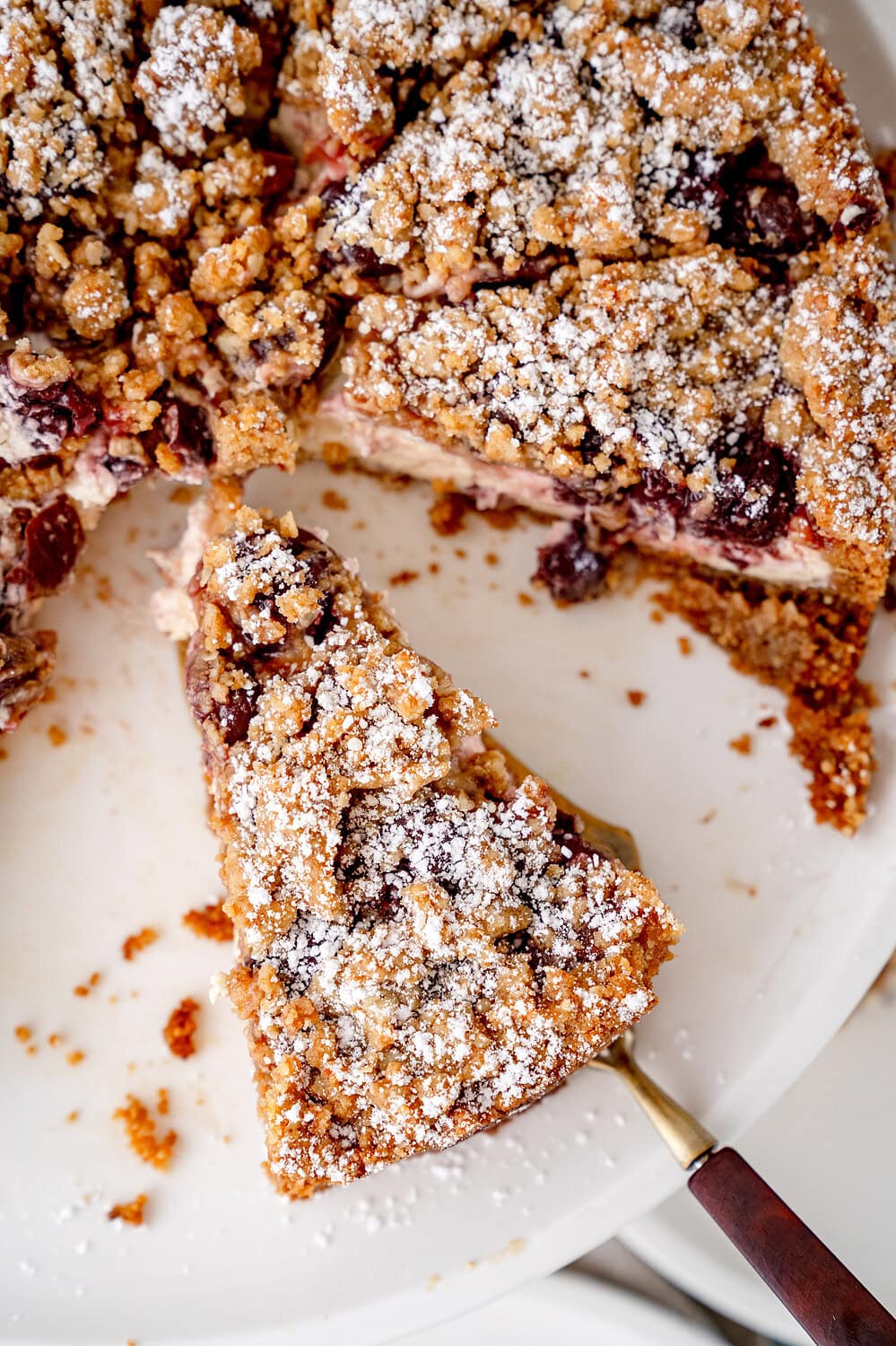 Topview auf einen Kirsch-streuselkuchen mit Vanillecreme und Keksboden, von dem gerade ein Stück auf dem Tortenheber liegt.